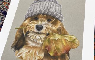 Print of dog holding a leaf in its mouth wearing a grey wooly hat